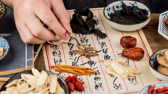 Practitioner's hand selecting Chinese herbs laid out over a handwritten herbal prescription